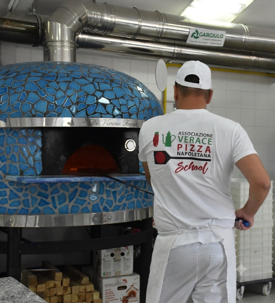 Pizzaiolo selin, valkoinen hattu ja paita, sininen kiviuuni ja savupiiput teollisuuskeittiössä.
